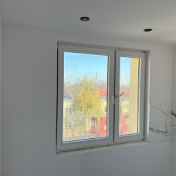 Jan Antoszczyk - Białe, świeżo pomalowane wnętrze z oknem na miasto. Widoczne punktowe oświetlenie sufitowe i fragment ściany wykończonej płytkami imitującymi marmur.