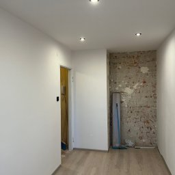 Jan Antoszczyk - Pokój po remoncie: białe ściany, drewniana podłoga, odsłonięta cegła z resztkami tynku. Widoczne punkty świetlne w suficie i fragment otwartych drzwi.