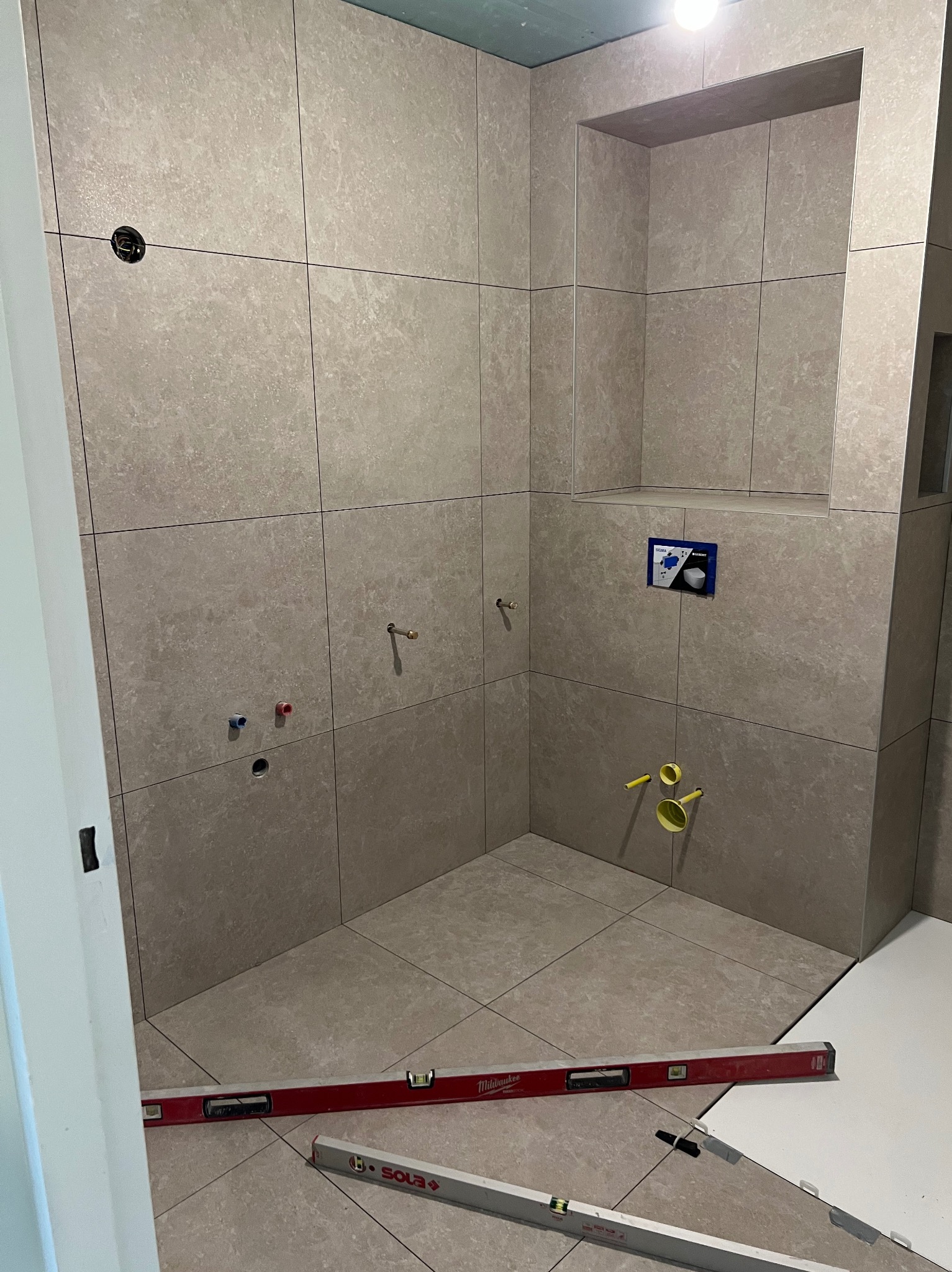 Beżowe płytki w łazience, częściowo ułożone. Widoczne poziomyce, instalacje wodne i wnęka w ścianie. Płytki ułożone równo, z zachowaniem fug.