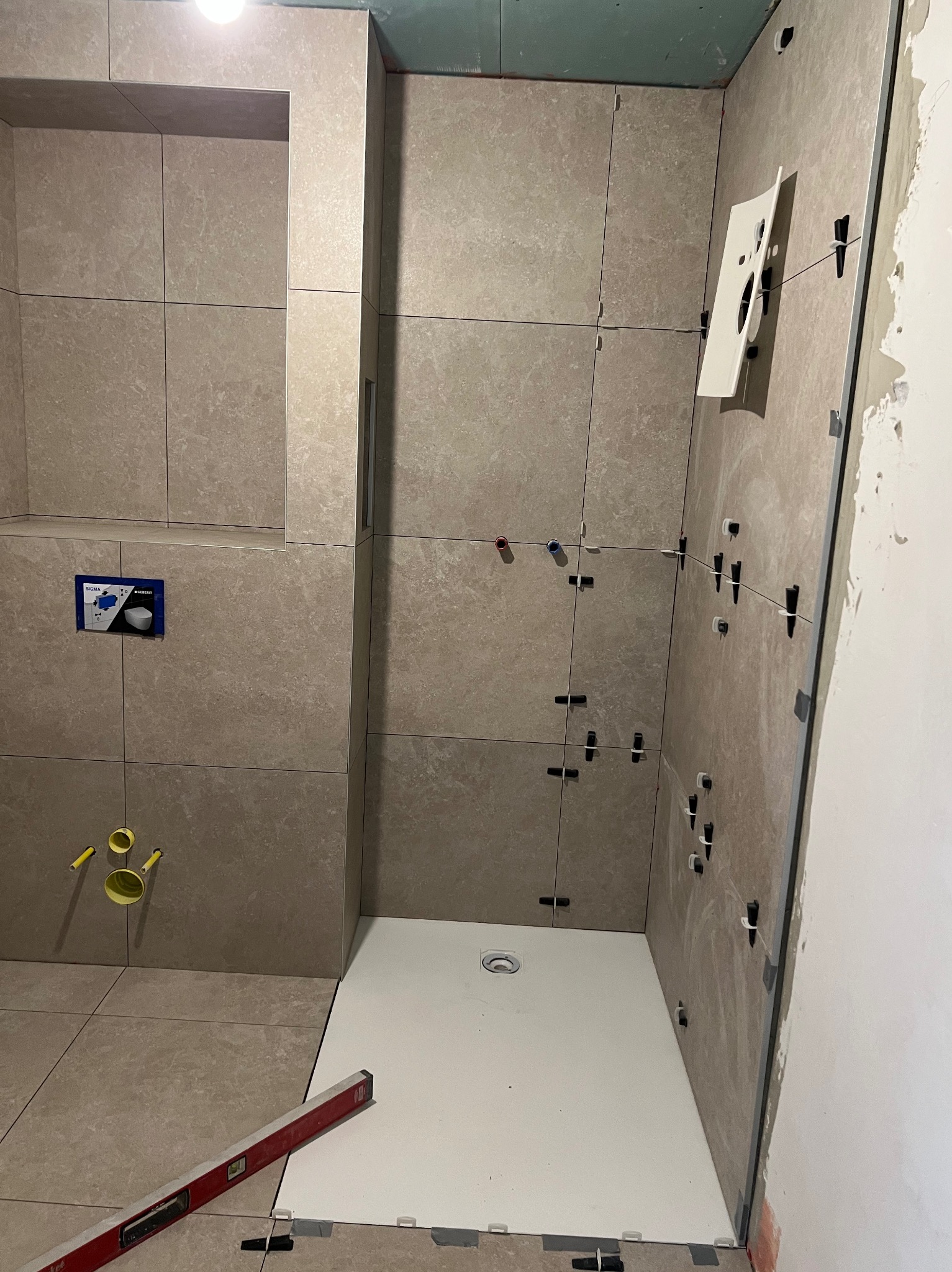 Montaż beżowych płytek w łazience z użyciem poziomicy i systemu poziomowania. Widoczny brodzik i fragment instalacji podtynkowej WC.