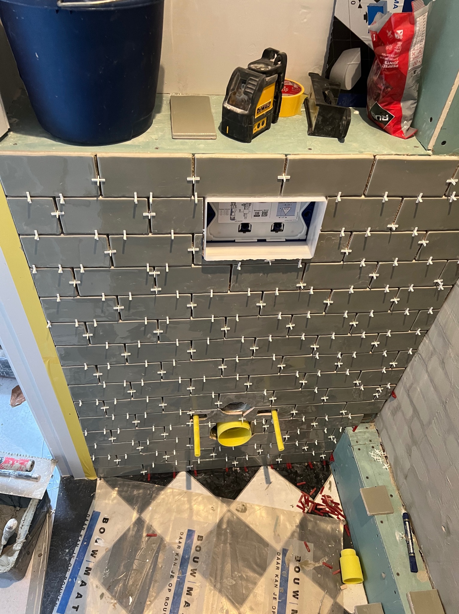 Układanie szarych płytek w łazience w Zduńskiej Woli. Widoczne krzyżyki dystansowe, poziomica laserowa i instalacja sanitarna. Prace glazurnicze w toku.