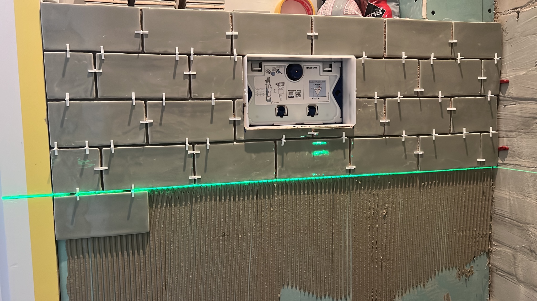 Układanie szarych płytek w łazience z użyciem poziomicy laserowej. Widoczna instalacja podtynkowa WC i krzyżyki dystansowe. Ściana pokryta klejem do płytek.