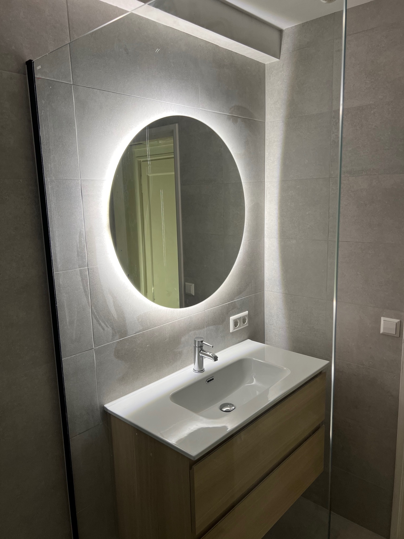 Szara łazienka z okrągłym lustrem LED, białą umywalką na drewnianej szafce i szklaną kabiną prysznicową. Minimalistyczny design, płytki imitujące kamień.