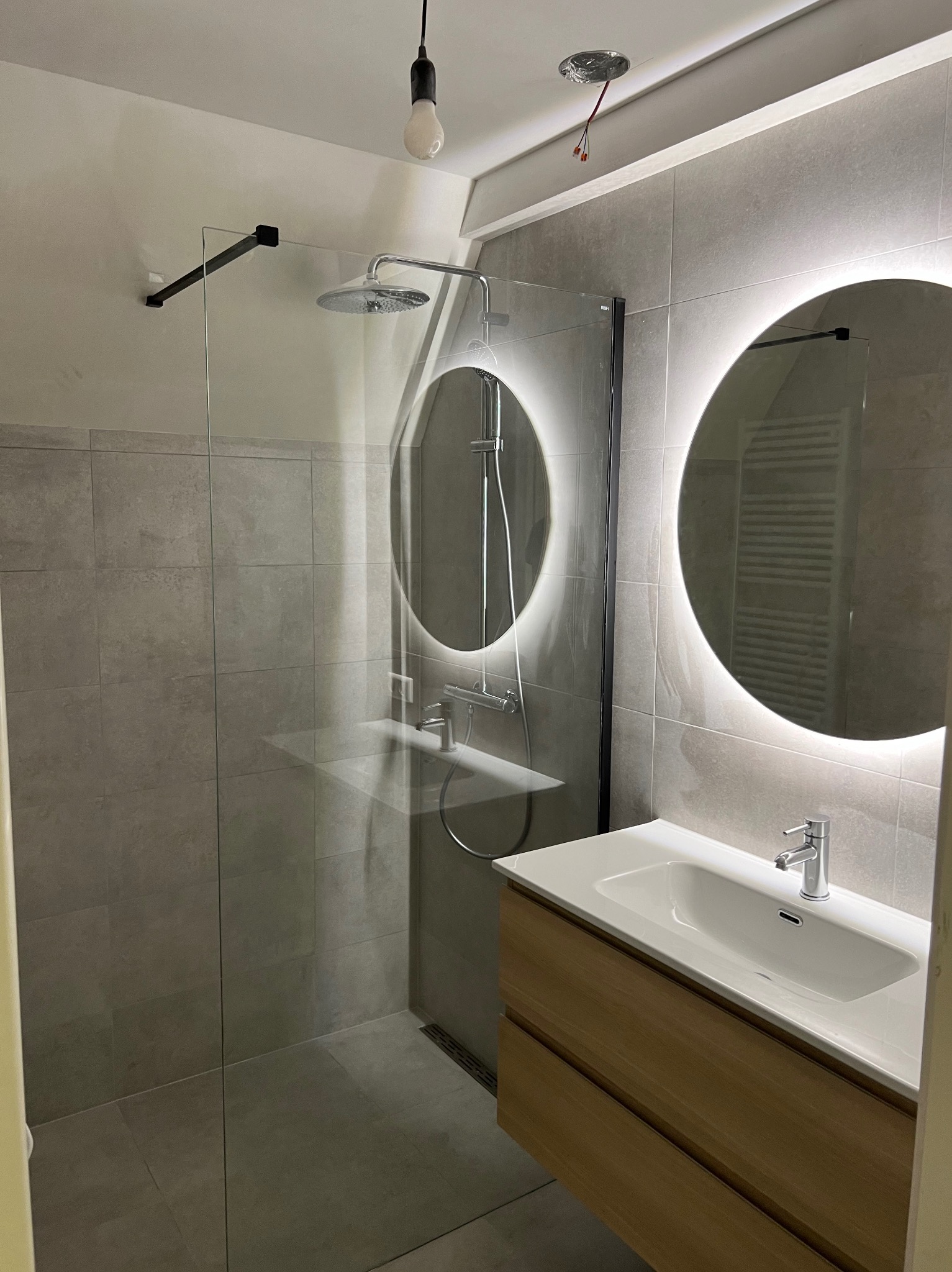 Nowoczesna łazienka z kabiną prysznicową typu walk-in, umywalką z szafką z jasnego drewna i okrągłymi lustrami z podświetleniem LED. Szare płytki na ścianach i podłodze.