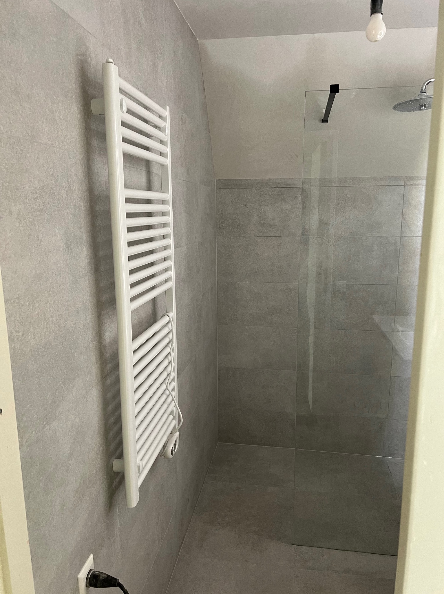 Nowoczesna łazienka z szarymi płytkami, biały grzejnik drabinkowy na ścianie, szklana kabina prysznicowa z czarnymi elementami i minimalistycznym oświetleniem.