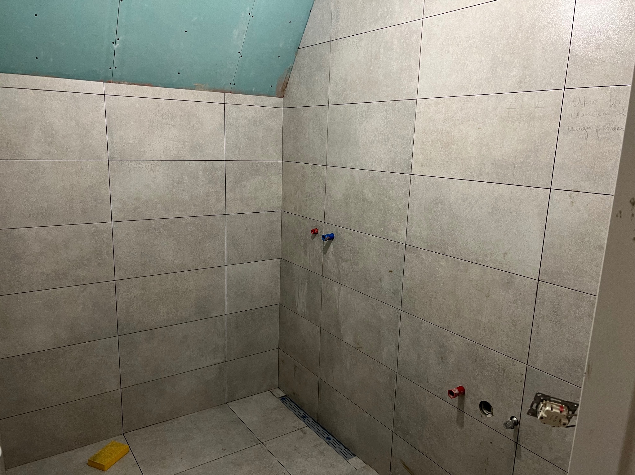 Łazienka w trakcie wykańczania: szare płytki na ścianach i podłodze, widoczne instalacje wodne i elektryczne, sufit częściowo wykończony płytą gipsową, odpływ liniowy.