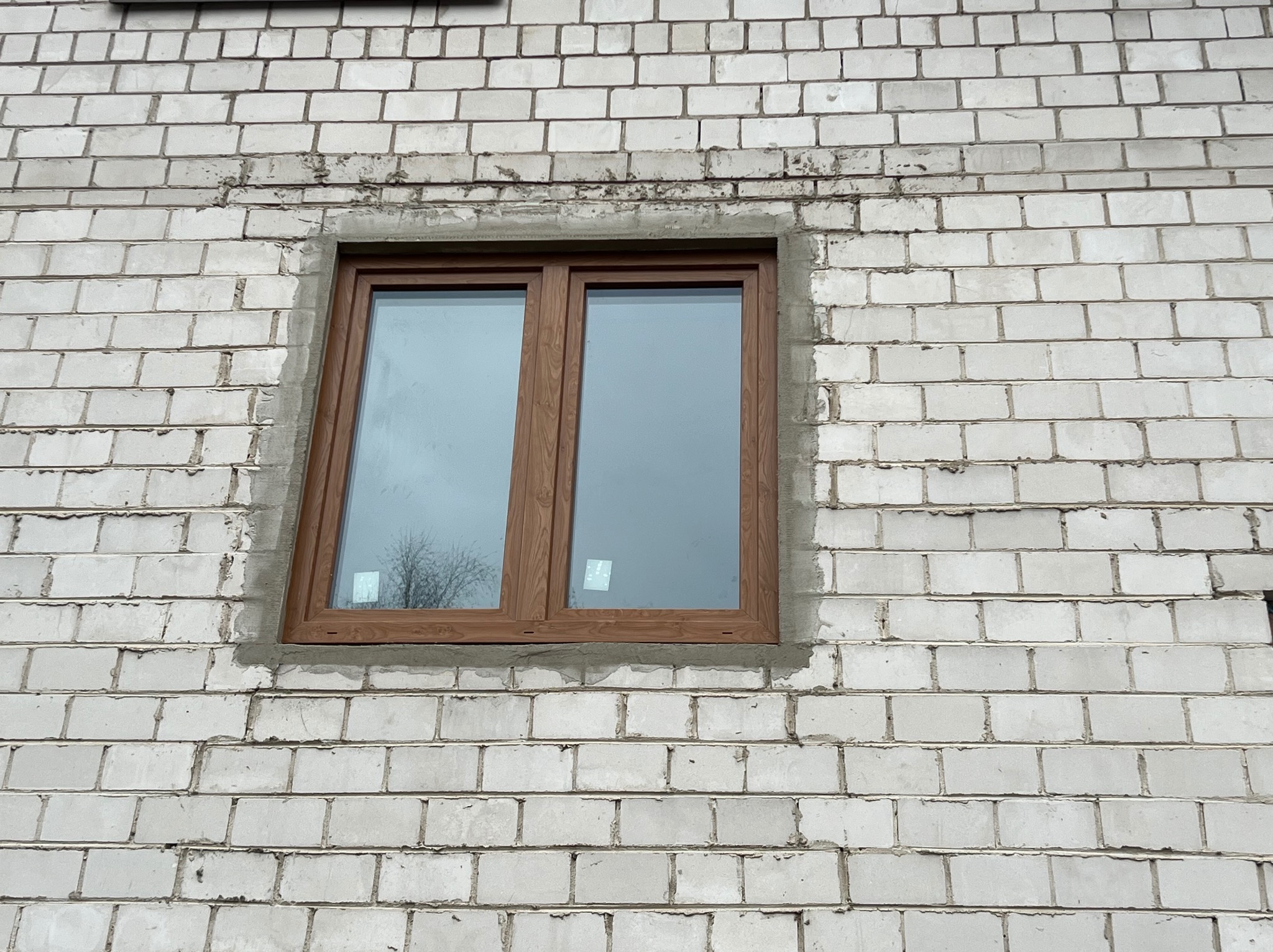 Nowe okno z brązową ramą w surowej, białej ścianie z pustaków. Widoczne ślady po montażu okna. W szybie odbija się drzewo.