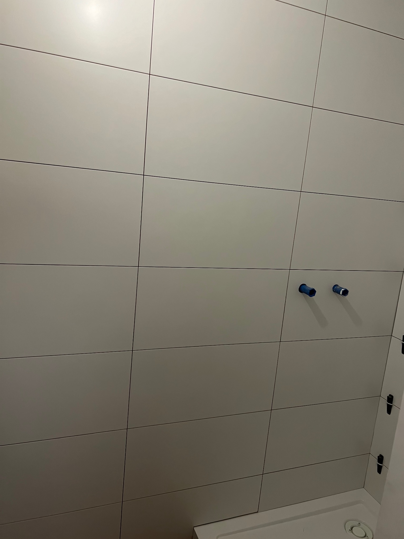 Ściana wyłożona jasnymi, kwadratowymi płytkami z ciemnymi fugami. Widoczne dwa niebieskie zakończenia rur. Na dole fragment brodzika.