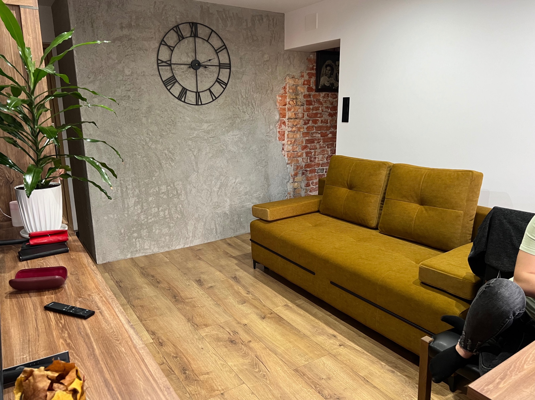 Nowoczesne wykończenie wnętrza: szara ściana z efektem betonu, zegar rzymski i fragment odsłoniętej cegły. Ustawiona na parkiecie musztardowa kanapa.