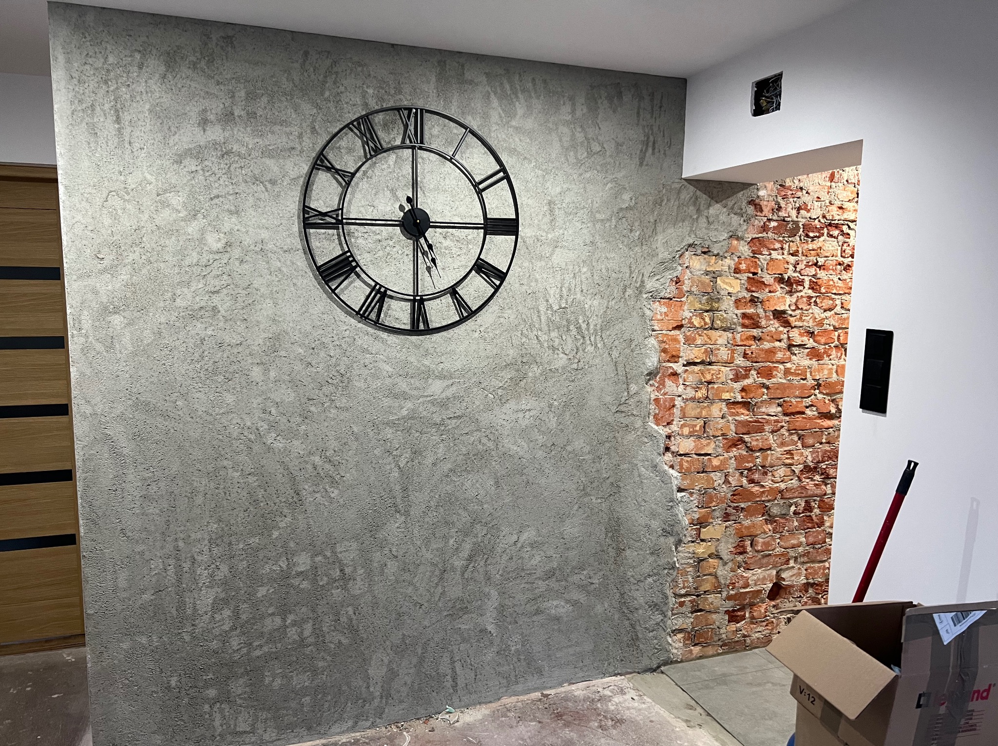 Ściana z dekoracyjną szarą strukturą i dużym zegarem, fragment odsłoniętej cegły przy przejściu. Widoczne kartony i narzędzie, sugerujące prace wykończeniowe.