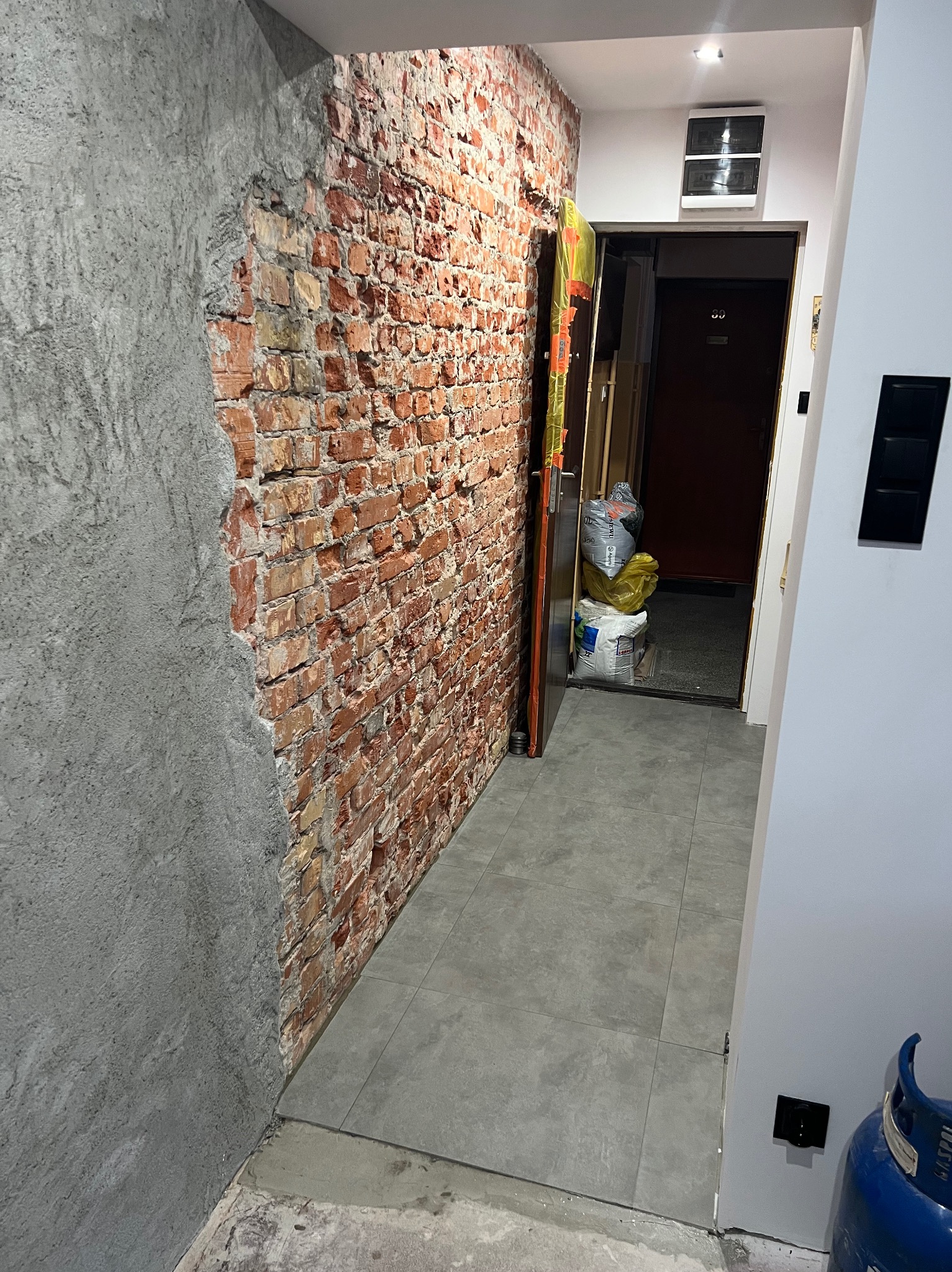 Korytarz w trakcie remontu: ściana z cegły częściowo pokryta gładzią, szare płytki na podłodze, worki z materiałami budowlanymi w przejściu, butla gazowa w rogu.
