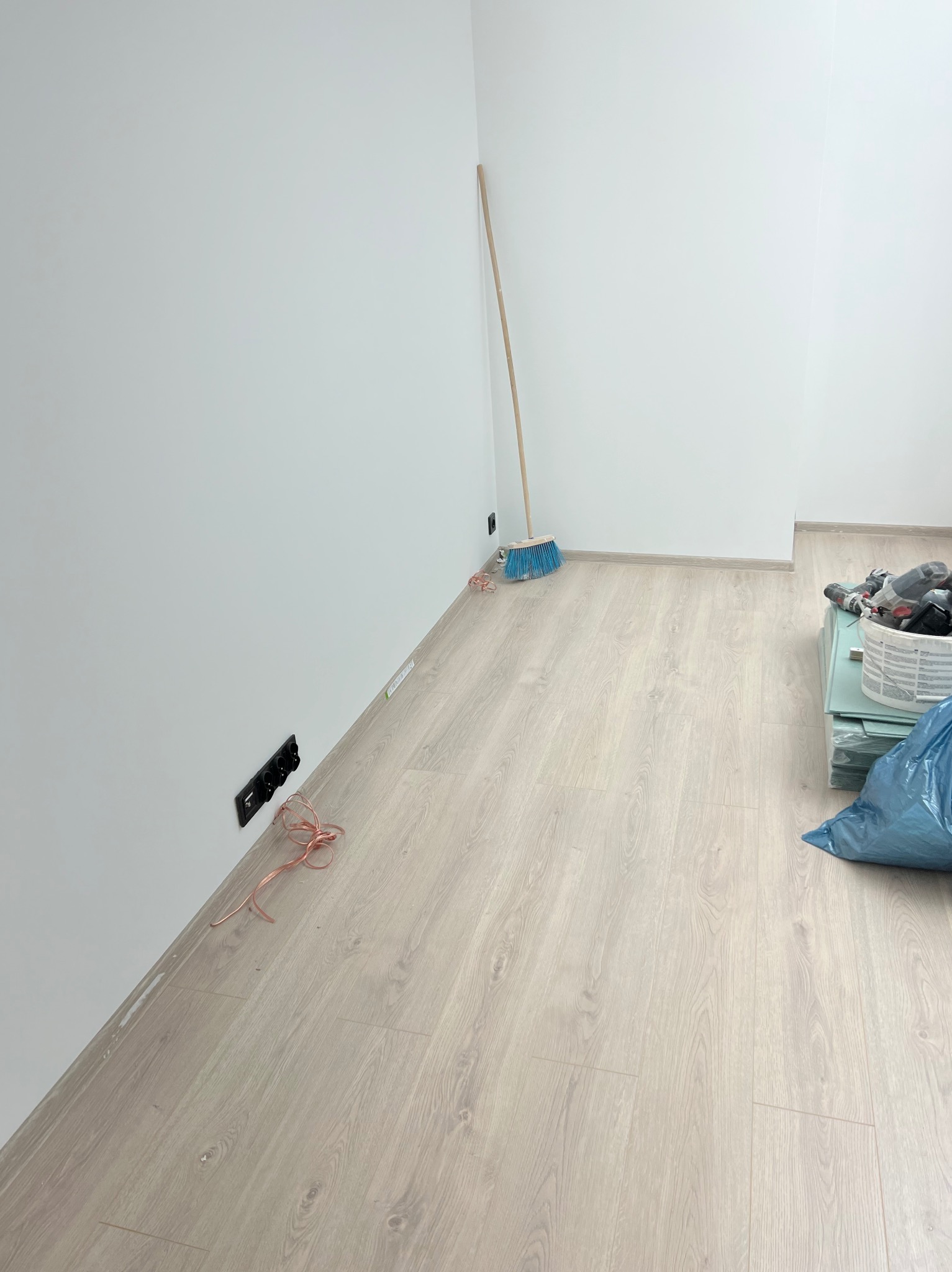 Pokój po remoncie: białe ściany, jasna podłoga, listwy przypodłogowe. Widoczna szczotka oparta o ścianę, gniazdka elektryczne i narzędzia w koszu.
