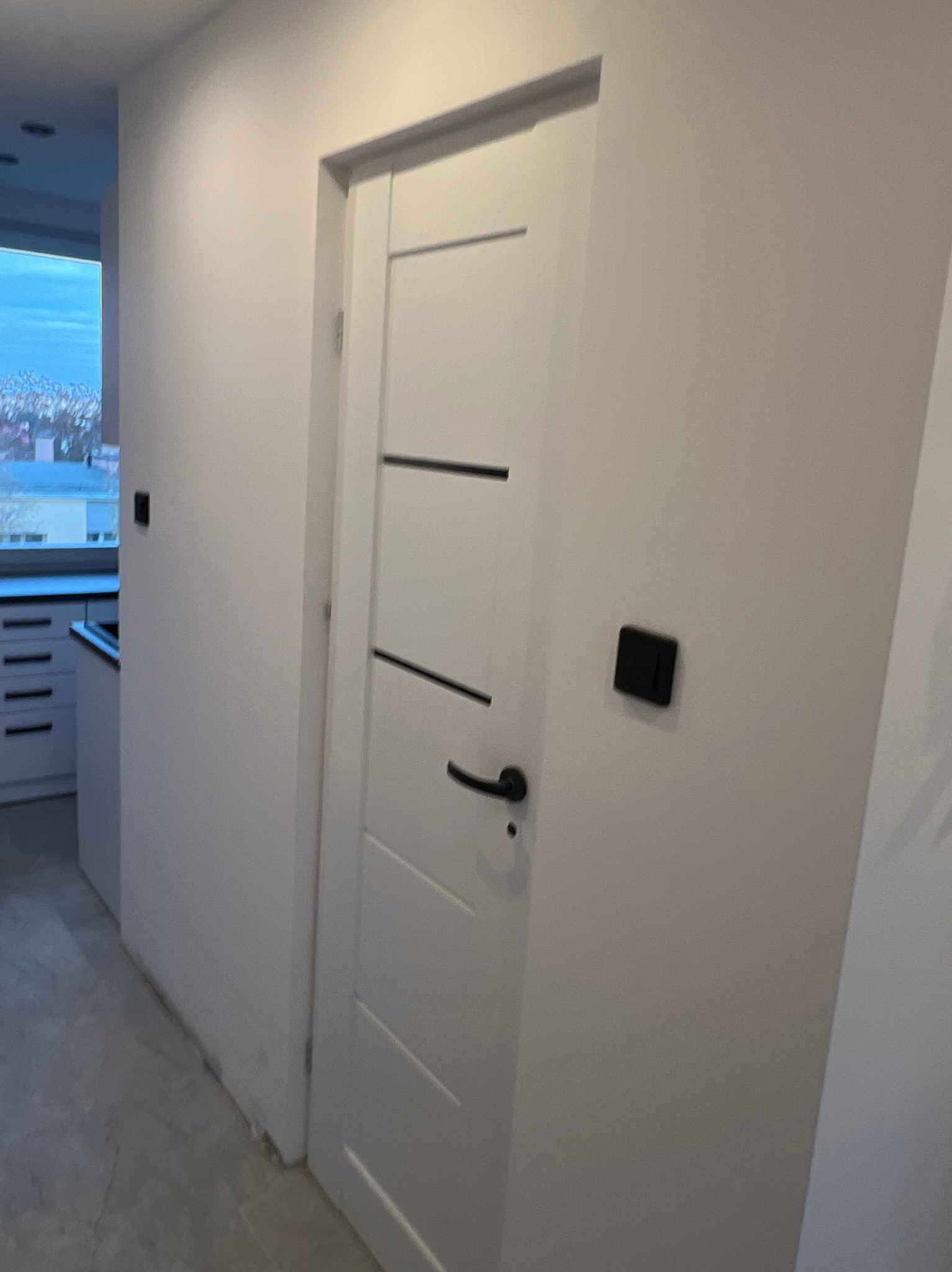 Wnętrze po remoncie: białe ściany, nowoczesne drzwi z czarnymi akcentami i włącznikiem. Widoczny fragment kuchni z białymi szafkami, okno z widokiem na miasto.