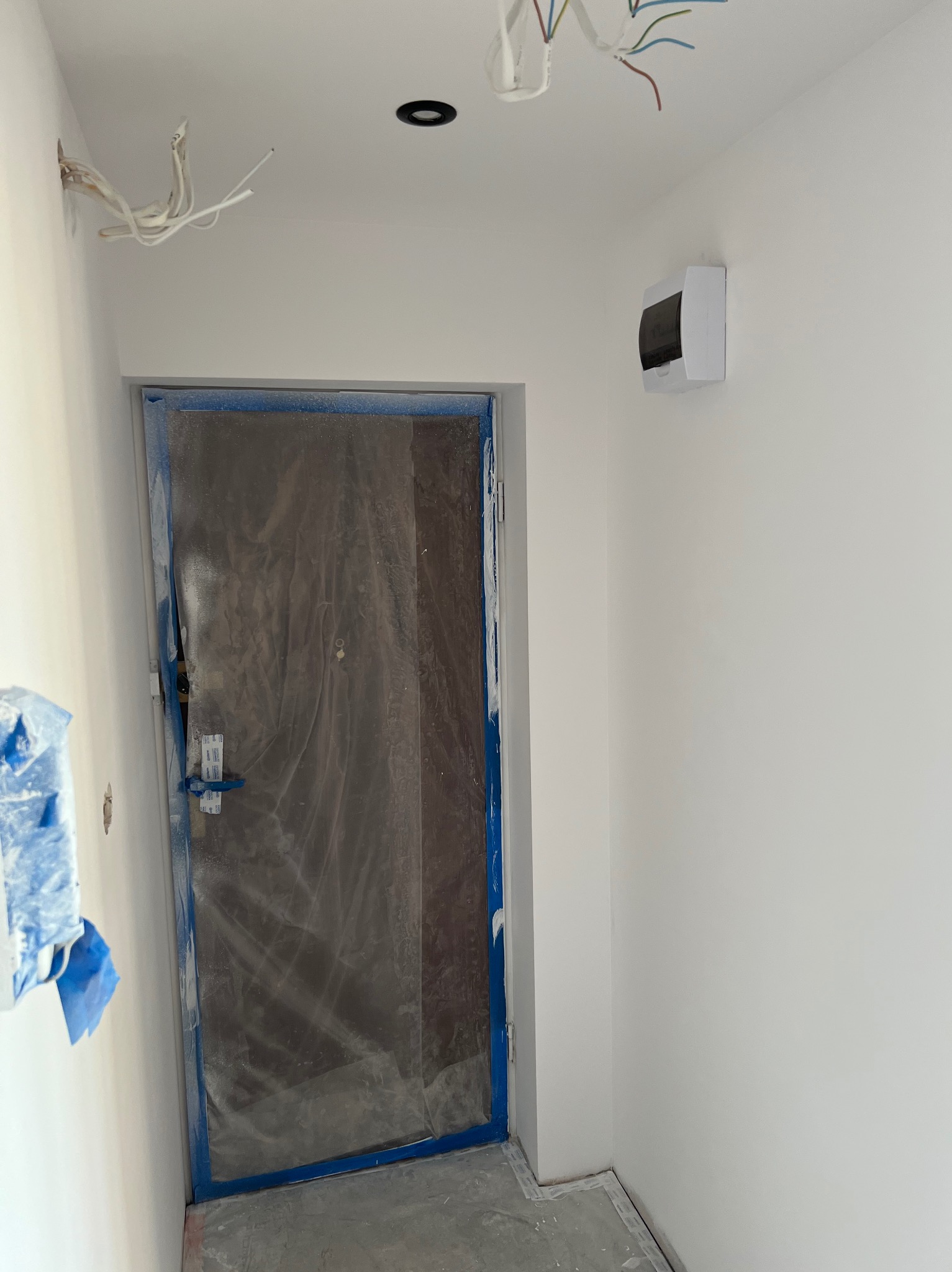 Białe ściany w trakcie malowania, zabezpieczone drzwi folią i taśmą malarską, widoczna instalacja elektryczna i skrzynka bezpiecznikowa.