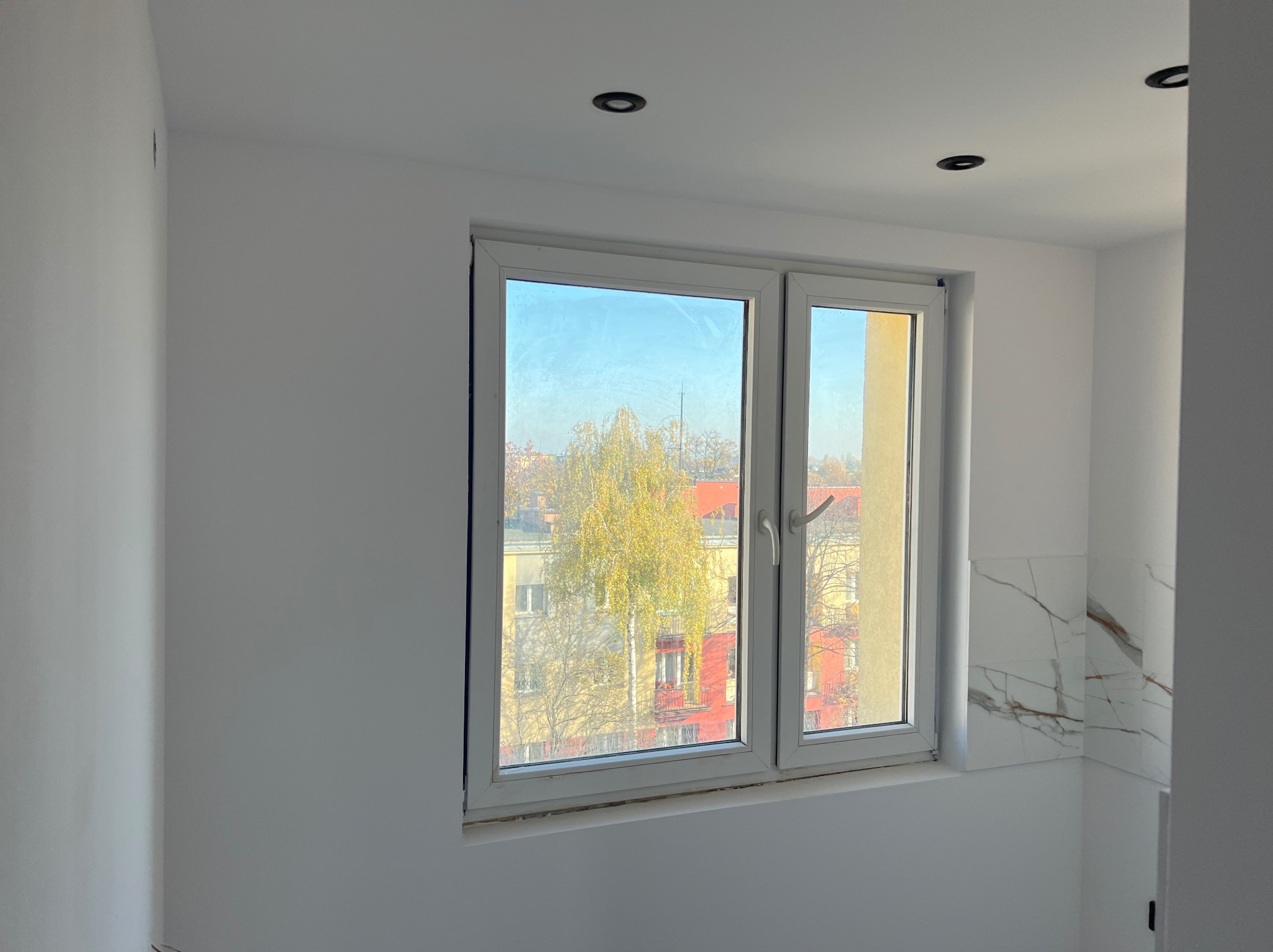 Białe, świeżo pomalowane wnętrze z oknem na miasto. Widoczne punktowe oświetlenie sufitowe i fragment ściany wykończonej płytkami imitującymi marmur.