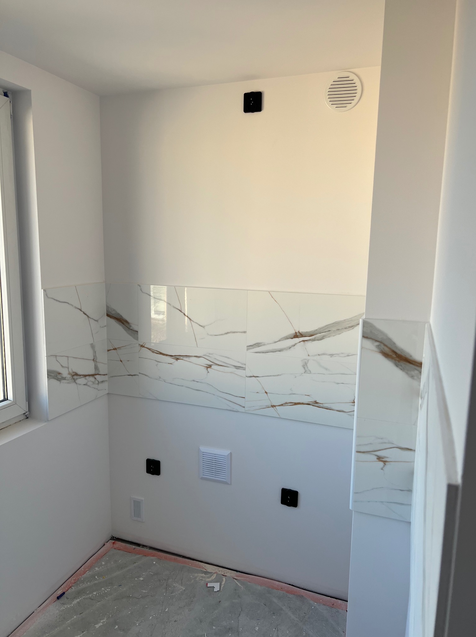 Wykończona łazienka z białymi płytkami imitującymi marmur na ścianach do połowy wysokości. Widoczne gniazdka elektryczne, wentylacja i okno. Podłoga w stanie surowym.