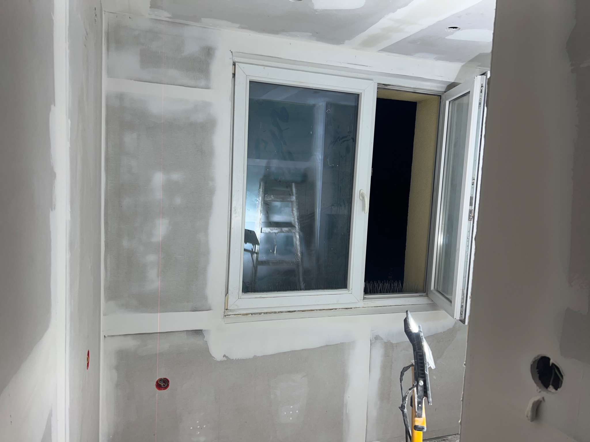 Pokój w trakcie prac wykończeniowych: ściany z płyt gipsowo-kartonowych, częściowo pokryte gładzią, okno otwarte, widoczna drabina i narzędzia na podłodze.