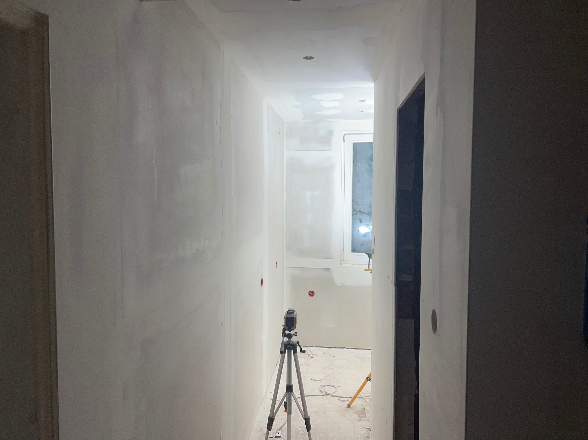 Korytarz w trakcie prac wykończeniowych. Ściany częściowo pokryte gładzią, widoczny laser krzyżowy na statywie, sufit z widocznymi punktami świetlnymi.