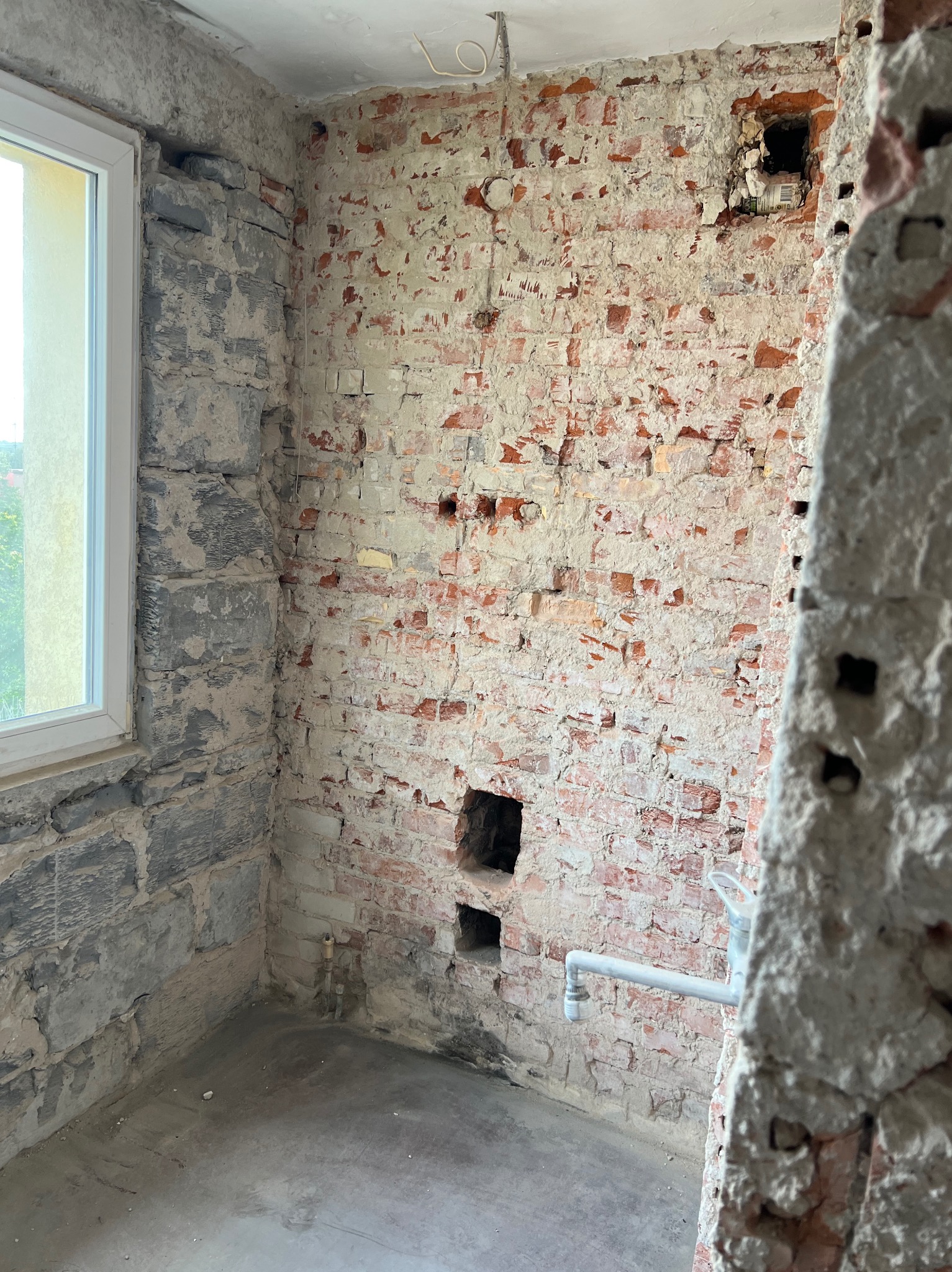 Ceglana ściana w trakcie prac remontowych, z widocznymi fragmentami starego tynku, otwory w ścianie i fragment instalacji wodnej. Okno z widokiem na zieleń.