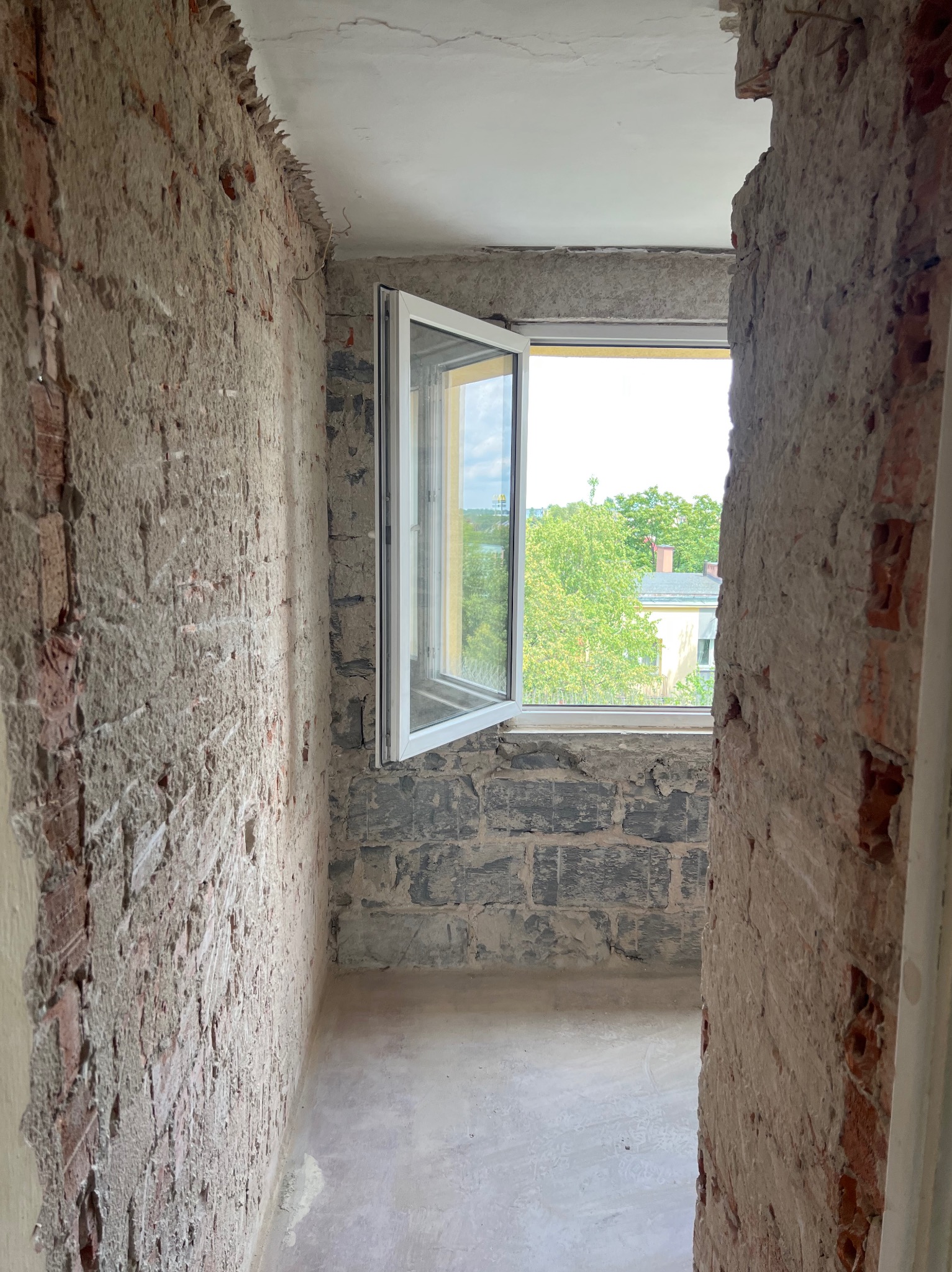 Wnętrze pokoju w trakcie remontu. Odsłonięta cegła na ścianach, otwarte okno z widokiem na zieleń. Widoczne ślady po starych instalacjach i przygotowania do dalszych prac wykończeniowych.