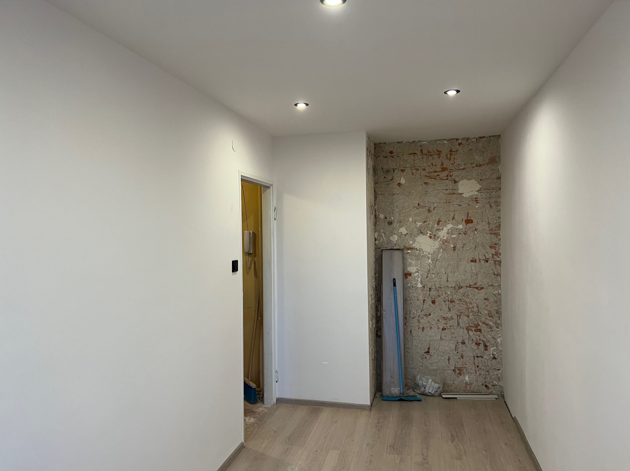 Pokój po remoncie: białe ściany, drewniana podłoga, odsłonięta cegła z resztkami tynku. Widoczne punkty świetlne w suficie i fragment otwartych drzwi.