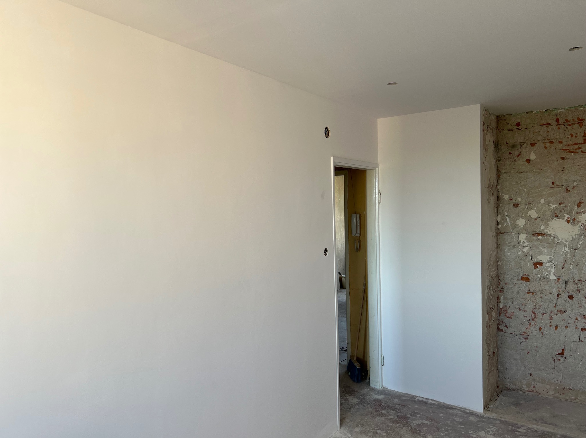 Wnętrze pokoju po remoncie: gładkie, białe ściany i sufit, fragment ściany z odsłoniętą cegłą, otwór drzwiowy. Widoczne ślady prac wykończeniowych na podłodze.