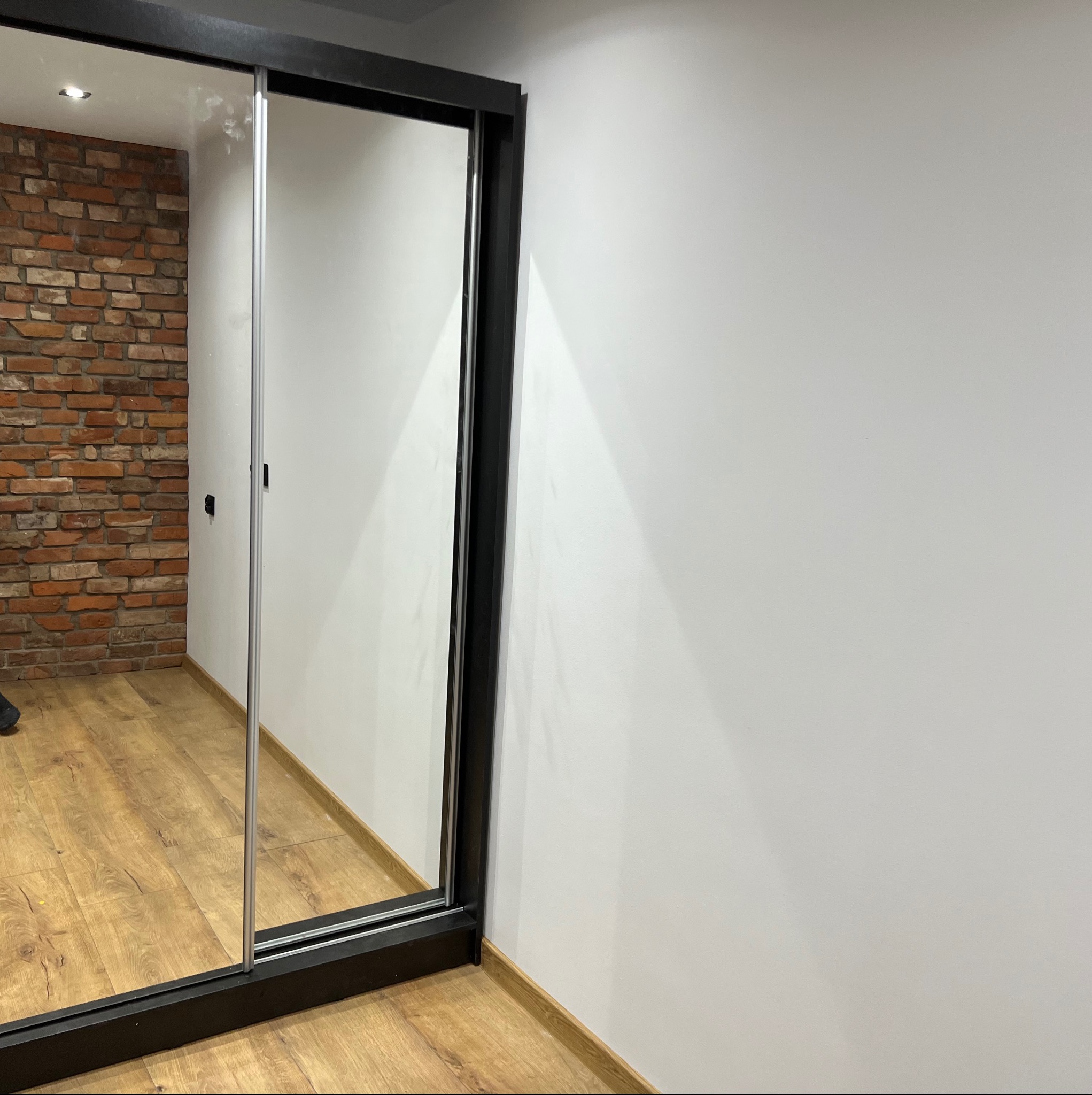 Wnętrze pokoju z przesuwną szafą z lustrem, odbijającą ceglaną ścianę i drewnianą podłogę. Minimalistyczny design z białą ścianą w tle.