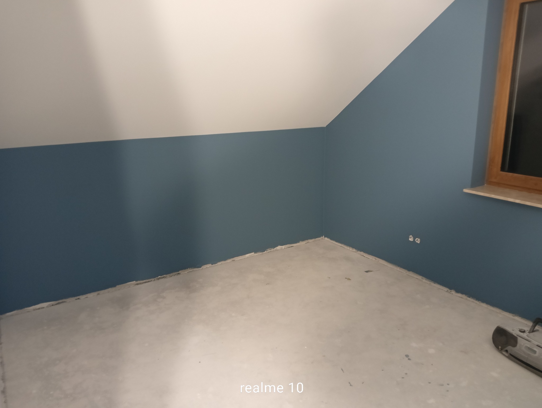 Pomieszczenie po malowaniu ścian na kolor niebieski, widoczne gniazdka elektryczne i drewniane okno. Podłoga surowa, przygotowana do dalszych prac wykończeniowych.