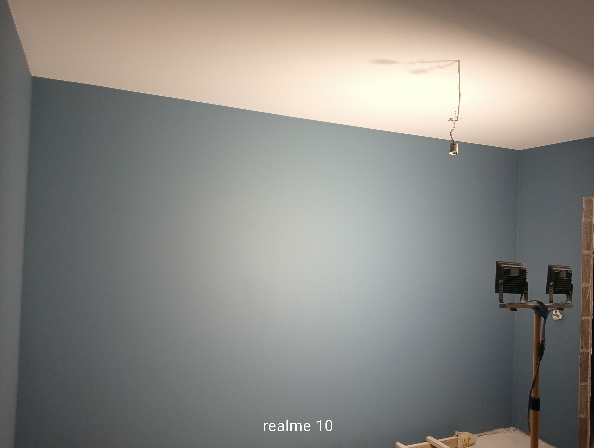 Ściana pomalowana na niebiesko-szary kolor, widoczne lampy budowlane i fragment instalacji elektrycznej. Świeżo pomalowane wnętrze w trakcie prac wykończeniowych.