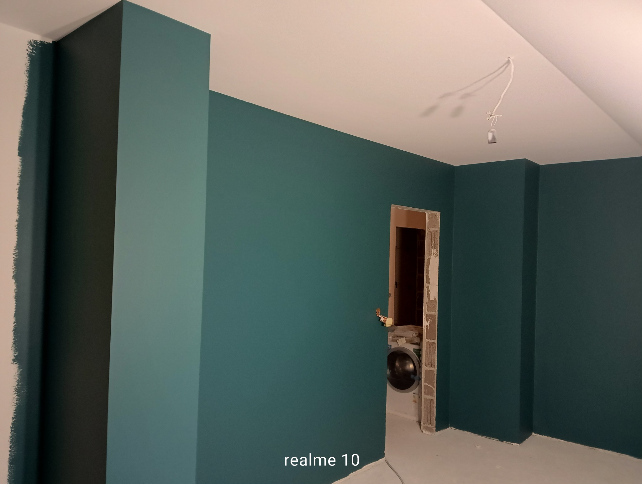 Pomieszczenie po malowaniu ścian na kolor morski. Widoczny fragment ściany w trakcie malowania, surowy otwór drzwiowy i wystające kable z sufitu.