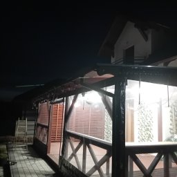 Dm - Zadaszona konstrukcja z drewnianą balustradą i przezroczystymi panelami bocznymi, oświetlona od wewnątrz, widoczna w nocnym otoczeniu z fragmentem budynku w tle.