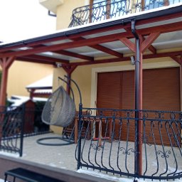 Dm - Zadaszony taras z drewnianą konstrukcją i balustradą z kutego żelaza, wiszące krzesło na tle budynku w zimowej scenerii.