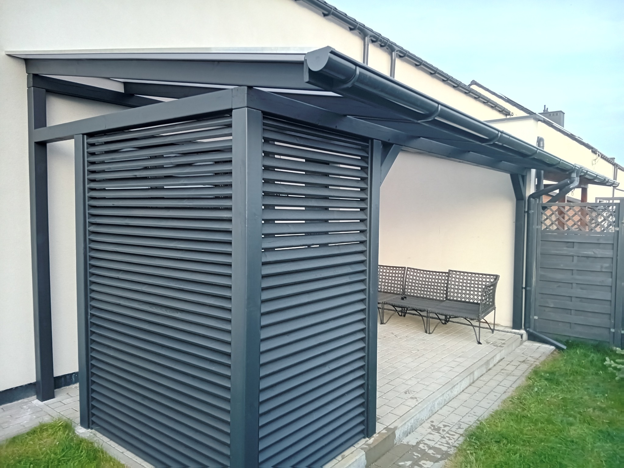 Nowoczesna pergola metalowa w kolorze antracytowym z żaluzjowymi panelami bocznymi, przylegająca do budynku z widocznym systemem rynnowym i ławką na wyłożonym kostką tarasie.