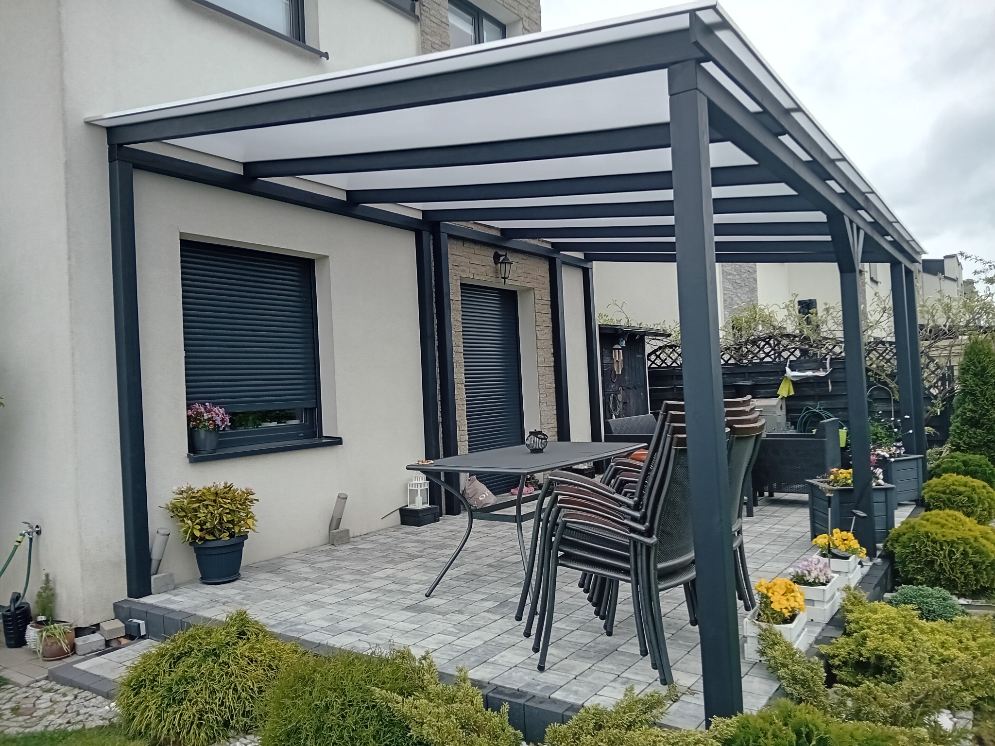 Nowoczesna pergola metalowa z białym zadaszeniem przylegająca do domu z szarymi roletami, na wybrukowanym tarasie stoi stół i krzesła, a całość otoczona jest zielenią.