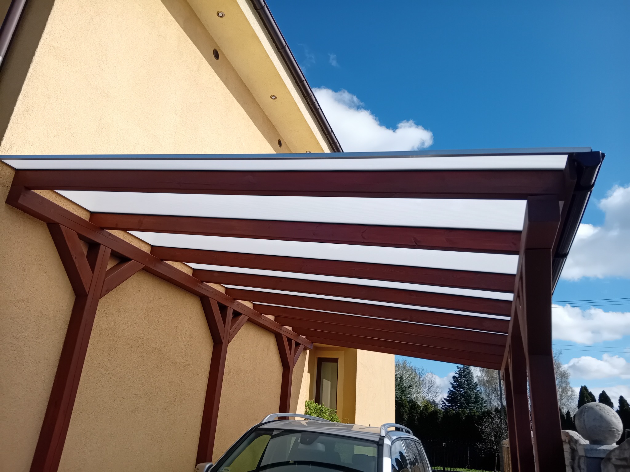Zadaszenie drewniane z białym, przezroczystym wypełnieniem, przylegające do ściany budynku, chroniące zaparkowany samochód przed słońcem i deszczem, z widocznym niebem w tle.