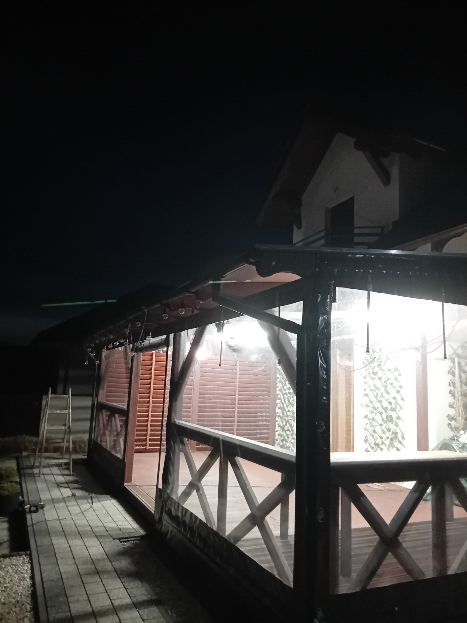 Zadaszona konstrukcja z drewnianą balustradą i przezroczystymi panelami bocznymi, oświetlona od wewnątrz, widoczna w nocnym otoczeniu z fragmentem budynku w tle.