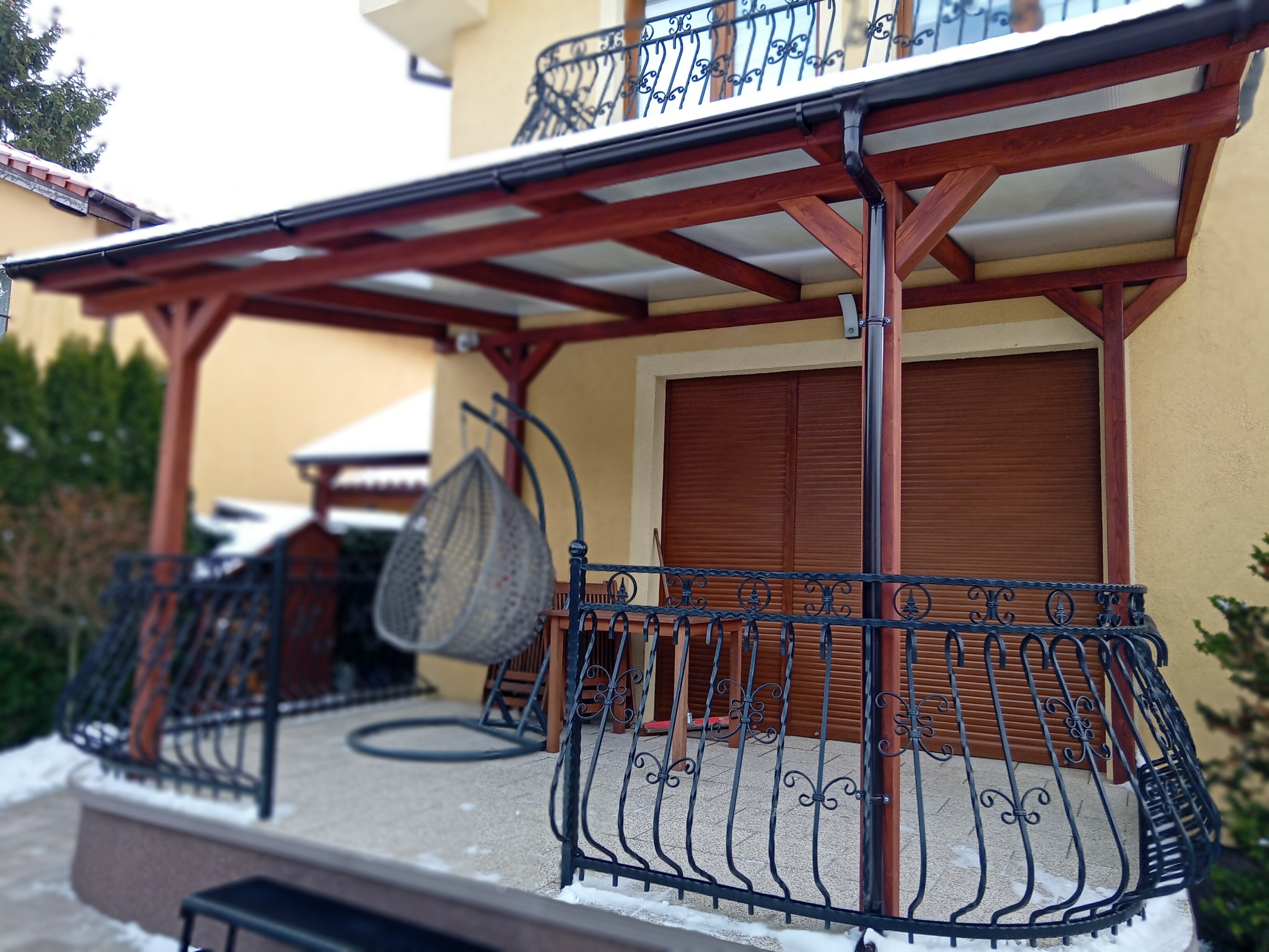 Zadaszony taras z drewnianą konstrukcją i balustradą z kutego żelaza, wiszące krzesło na tle budynku w zimowej scenerii.