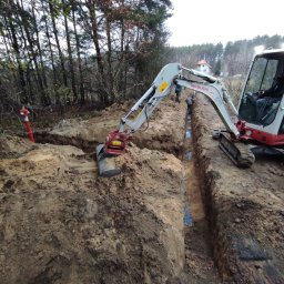 HYDRO-DUO Sp. z o. o. - Wykonanie wykopu pod rury niebieskie, widoczny mały traktor Takeuchi. W tle las i budynek. Czerwony hydrant po lewej. Prace ziemne w toku, widoczne osoby w oddali.