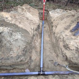 HYDRO-DUO Sp. z o. o. - Ziemny wykop z niebieskimi rurami wodociągowymi i czerwonym hydrantem na tle leśnego krajobrazu. Widoczne narzędzia: łopata i grabie. Ujęcie z góry.