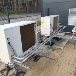 HYDRO-DUO Sp. z o. o. - Dwie białe pompy ciepła zamontowane na zewnątrz budynku z szarą elewacją. Widoczne rury i elementy instalacji, tło stanowią drzewa i ogrodzenie.