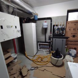 HYDRO-DUO Sp. z o. o. - Zmontowana instalacja grzewcza z pompą ciepła Eco Wave, zbiornikami i rozdzielaczem w pomieszczeniu z szarą glazurą i kartonami na podłodze. Widoczne rury i przewody.