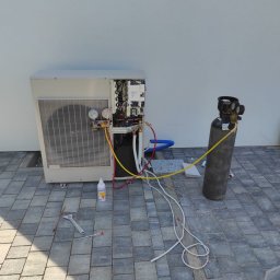 HYDRO-DUO Sp. z o. o. - Serwis pompy ciepła: otwarty panel, manometry, butla z gazem chłodniczym, narzędzia na kostce brukowej. Prace serwisowe w toku, widoczne przewody i elementy instalacji.