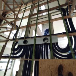 HYDRO-DUO Sp. z o. o. - Montaż instalacji CO w surowym budynku: czarne, karbowane rury prowadzone pod stropem, mocowane do konstrukcji drewnianej. Widoczne okna i fragment podłogi.