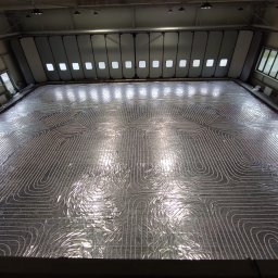 HYDRO-DUO Sp. z o. o. - Błyszcząca, aluminiowa folia z ułożonymi rurami ogrzewania podłogowego w dużym pomieszczeniu. Widok z góry ukazuje spiralny wzór instalacji.
