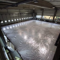 HYDRO-DUO Sp. z o. o. - Rozległa instalacja ogrzewania podłogowego z białych rur ułożonych na srebrnej folii izolacyjnej w dużej hali przemysłowej, widok z góry.
