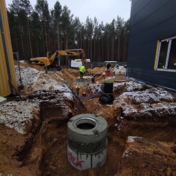 HYDRO-DUO Sp. z o. o. - Montaż studni betonowej w wykopie, widoczny pracownik w kamizelce, koparka CAT w tle, częściowo ośnieżona ziemia i budynek w tle. Prace ziemne.