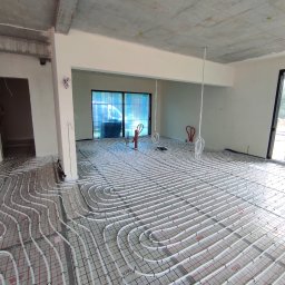 HYDRO-DUO Sp. z o. o. - Białe rury ogrzewania podłogowego, ułożone na izolacji z siatką, w surowym wnętrzu z betonowym sufitem i dużymi oknami. Widok na rozległą instalację przed zalaniem posadzki.