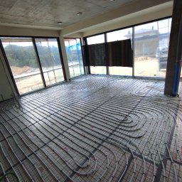 HYDRO-DUO Sp. z o. o. - Instalacja ogrzewania podłogowego w surowym wnętrzu z dużymi oknami. Rury grzewcze ułożone na specjalnej siatce, widoczne złącza i fragmenty instalacji.