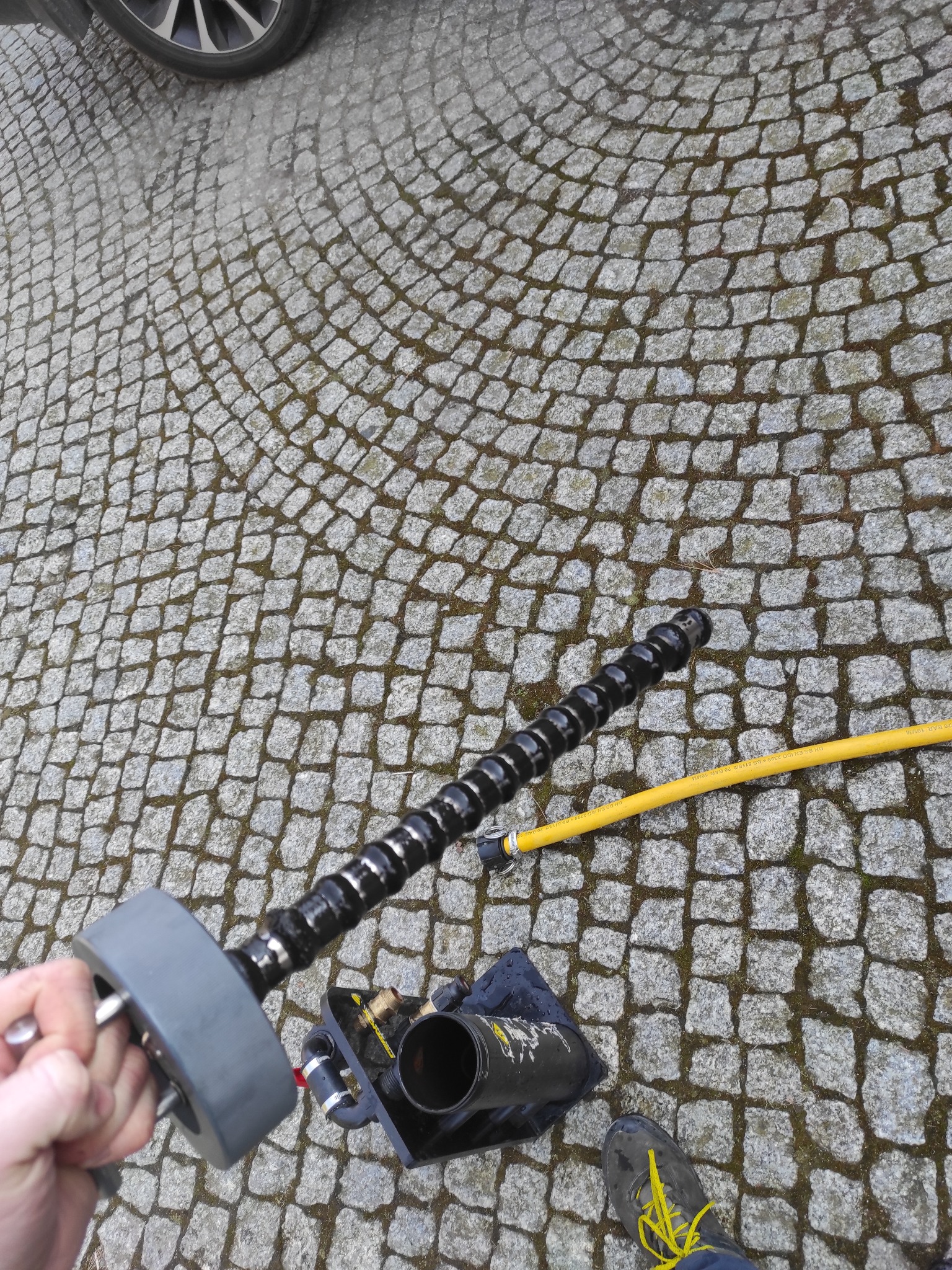 Czyszczenie drenażu przydomowej oczyszczalni ścieków za pomocą spirali, widoczna studzienka, żółty wąż i fragment buta na kostce brukowej.