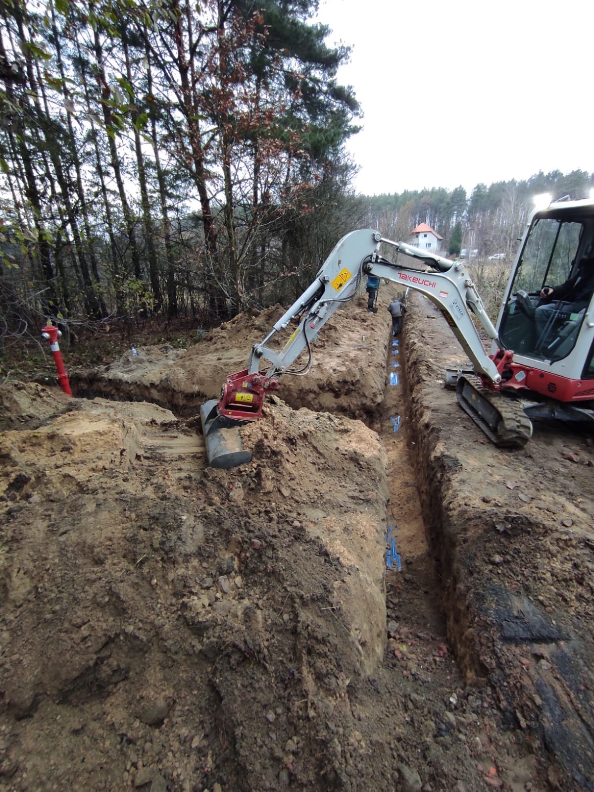 Wykonanie wykopu pod rury niebieskie, widoczny mały traktor Takeuchi. W tle las i budynek. Czerwony hydrant po lewej. Prace ziemne w toku, widoczne osoby w oddali.